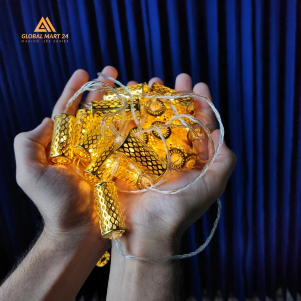 A pair of hands is holding a glowing string of decorative golden LED lights with cylindrical metal covers featuring a mesh design. The lights emit a warm yellow glow, and the background is a deep blue curtain. The logo of "GLOBAL MART 24" is visible in the upper left corner with the tagline "MAKING LIFE EASIER."