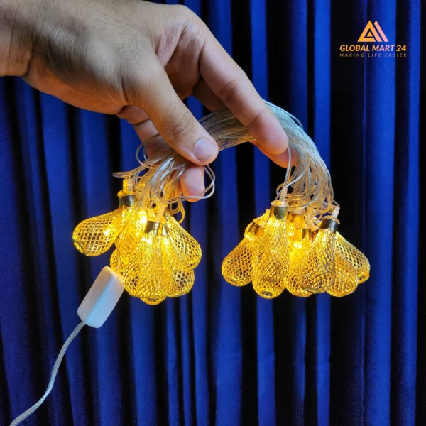 A pair of hands is holding a glowing string of decorative golden LED lights with cylindrical metal covers featuring a mesh design. The lights emit a warm yellow glow, and the background is a deep blue curtain. The logo of "GLOBAL MART 24" is visible in the upper left corner with the tagline "MAKING LIFE EASIER."