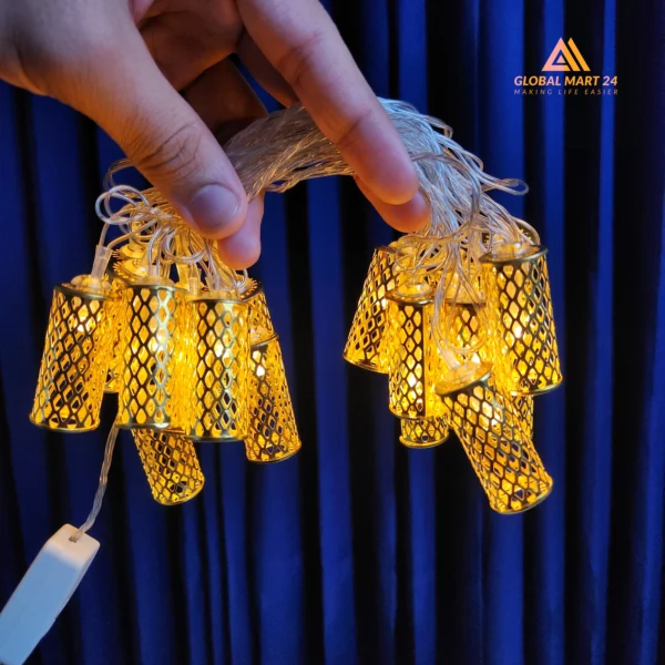 A pair of hands is holding a glowing string of decorative golden LED lights with cylindrical metal covers featuring a mesh design. The lights emit a warm yellow glow, and the background is a deep blue curtain. The logo of "GLOBAL MART 24" is visible in the upper left corner with the tagline "MAKING LIFE EASIER."