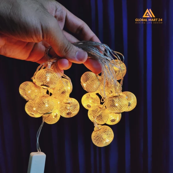 A pair of hands is holding a glowing string of decorative golden LED lights with cylindrical metal covers featuring a mesh design. The lights emit a warm yellow glow, and the background is a deep blue curtain. The logo of "GLOBAL MART 24" is visible in the upper left corner with the tagline "MAKING LIFE EASIER."