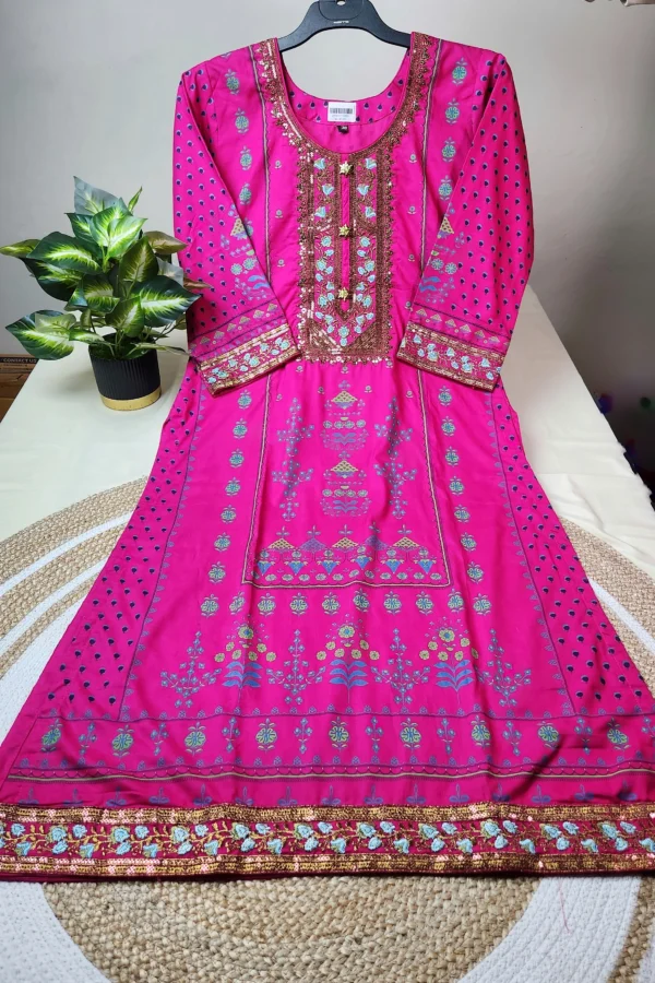 "Bright pink traditional South Asian kurti displayed on a hanger, adorned with intricate embroidery in gold, turquoise, and blue. The neckline features heavy sequin and thread work in a rectangular pattern, while the sleeves and hemline are decorated with matching ornate designs. The kurti has floral and geometric motifs throughout and is laid out neatly on a white surface next to a potted plant."