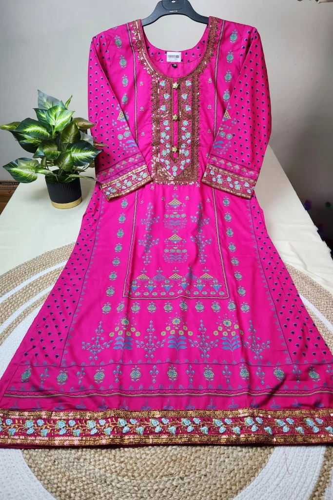 "Bright pink traditional South Asian kurti displayed on a hanger, adorned with intricate embroidery in gold, turquoise, and blue. The neckline features heavy sequin and thread work in a rectangular pattern, while the sleeves and hemline are decorated with matching ornate designs. The kurti has floral and geometric motifs throughout and is laid out neatly on a white surface next to a potted plant."