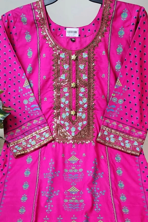 "Bright pink traditional South Asian kurti displayed on a hanger, adorned with intricate embroidery in gold, turquoise, and blue. The neckline features heavy sequin and thread work in a rectangular pattern, while the sleeves and hemline are decorated with matching ornate designs. The kurti has floral and geometric motifs throughout and is laid out neatly on a white surface next to a potted plant."