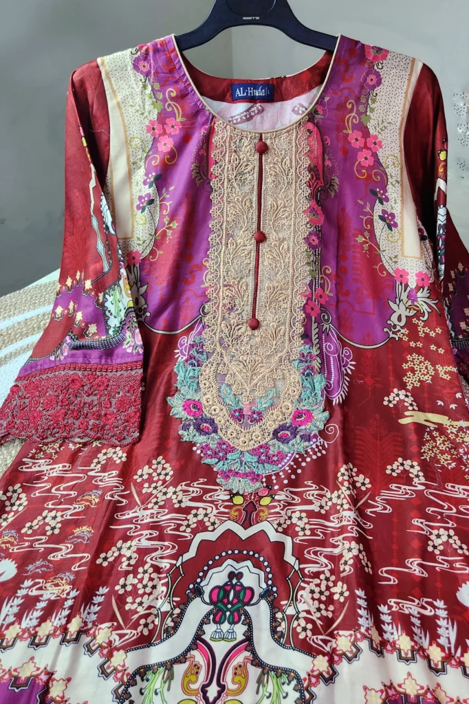 A vibrant traditional South Asian kurti in deep red and magenta tones with elaborate multicolor patterns and embroidery. The neckline features beige and gold lace embroidery accented with turquoise, pink, and purple floral threadwork, along with decorative red buttons down the center. The kurti showcases printed motifs of flowers, waves, and ornamental designs in cream, white, and various bright shades. The sleeves are adorned with intricate red lace embroidery. The garment is displayed on a black hanger against a neutral background.