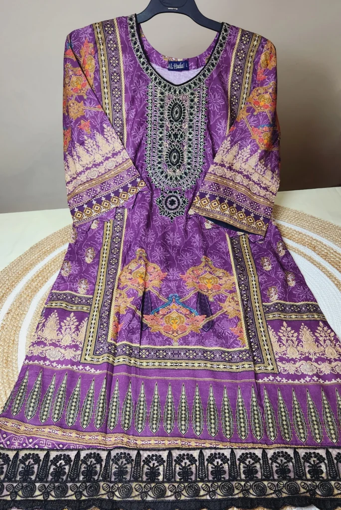 A traditional South Asian long kurti in rich purple with intricate multicolored patterns and embroidery. The neckline is heavily embroidered with black, silver, and green thread in an ornate design. The fabric features printed floral and geometric motifs in gold, orange, pink, and black. The sleeves and hem are bordered with elaborate designs, including leaf-like and floral patterns. The kurti is displayed on a black hanger, laid out neatly on a woven mat.