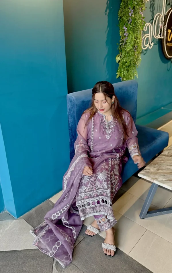 A woman stands indoors next to a bright blue wall under a wall-mounted light, wearing a traditional South Asian outfit. Her ensemble includes a soft purple kameez adorned with intricate white embroidery, paired with matching trousers and a sheer dupatta featuring circular and floral white patterns. She has long brown hair styled down and wears minimal jewelry. The setting includes a blue velvet bench, a marble-top table, and a vertical plant wall with purple flowers and decorative neon signage.