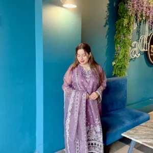 A woman stands indoors next to a bright blue wall under a wall-mounted light, wearing a traditional South Asian outfit. Her ensemble includes a soft purple kameez adorned with intricate white embroidery, paired with matching trousers and a sheer dupatta featuring circular and floral white patterns. She has long brown hair styled down and wears minimal jewelry. The setting includes a blue velvet bench, a marble-top table, and a vertical plant wall with purple flowers and decorative neon signage.