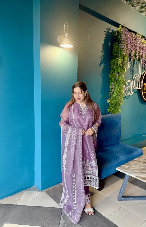 A woman stands indoors next to a bright blue wall under a wall-mounted light, wearing a traditional South Asian outfit. Her ensemble includes a soft purple kameez adorned with intricate white embroidery, paired with matching trousers and a sheer dupatta featuring circular and floral white patterns. She has long brown hair styled down and wears minimal jewelry. The setting includes a blue velvet bench, a marble-top table, and a vertical plant wall with purple flowers and decorative neon signage.