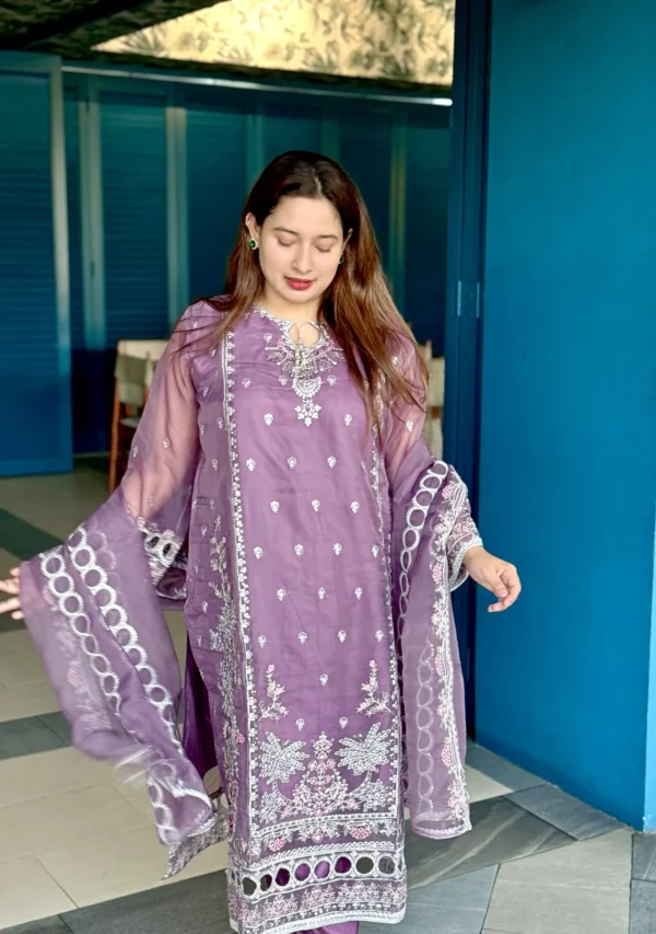 A woman stands indoors next to a bright blue wall under a wall-mounted light, wearing a traditional South Asian outfit. Her ensemble includes a soft purple kameez adorned with intricate white embroidery, paired with matching trousers and a sheer dupatta featuring circular and floral white patterns. She has long brown hair styled down and wears minimal jewelry. The setting includes a blue velvet bench, a marble-top table, and a vertical plant wall with purple flowers and decorative neon signage.