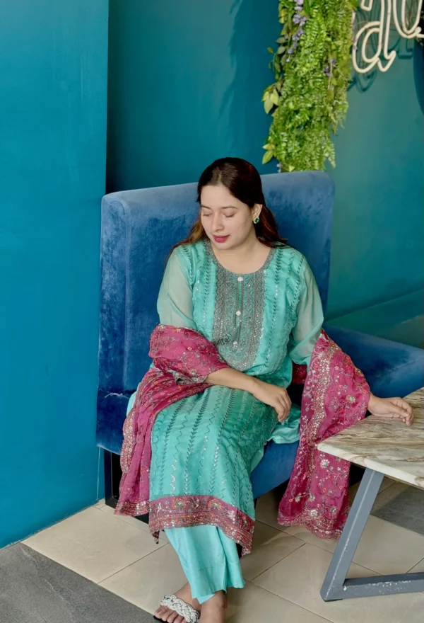 A woman is standing indoors against a bright blue wall, wearing a traditional South Asian outfit. Her dress is a turquoise blue kameez with intricate silver embroidery, paired with matching trousers. She holds a contrasting magenta dupatta with floral embroidery and sequin detailing. Her long brown hair is styled straight with a flower tucked behind her ear. The setting includes a modern interior with tiled flooring, wooden chairs, and a large window with greenery outside.