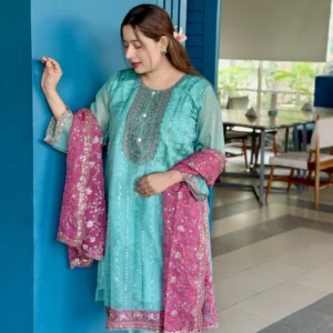 A woman is standing indoors against a bright blue wall, wearing a traditional South Asian outfit. Her dress is a turquoise blue kameez with intricate silver embroidery, paired with matching trousers. She holds a contrasting magenta dupatta with floral embroidery and sequin detailing. Her long brown hair is styled straight with a flower tucked behind her ear. The setting includes a modern interior with tiled flooring, wooden chairs, and a large window with greenery outside.