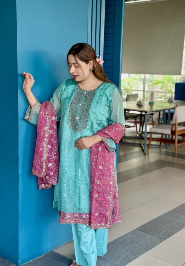 A woman is standing indoors against a bright blue wall, wearing a traditional South Asian outfit. Her dress is a turquoise blue kameez with intricate silver embroidery, paired with matching trousers. She holds a contrasting magenta dupatta with floral embroidery and sequin detailing. Her long brown hair is styled straight with a flower tucked behind her ear. The setting includes a modern interior with tiled flooring, wooden chairs, and a large window with greenery outside.