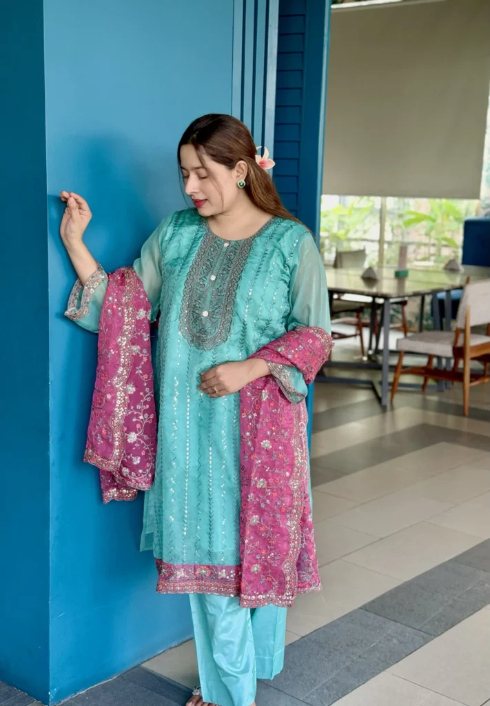 A woman is standing indoors against a bright blue wall, wearing a traditional South Asian outfit. Her dress is a turquoise blue kameez with intricate silver embroidery, paired with matching trousers. She holds a contrasting magenta dupatta with floral embroidery and sequin detailing. Her long brown hair is styled straight with a flower tucked behind her ear. The setting includes a modern interior with tiled flooring, wooden chairs, and a large window with greenery outside.