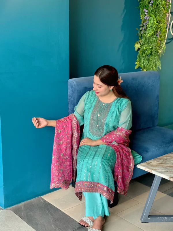 A woman is standing indoors against a bright blue wall, wearing a traditional South Asian outfit. Her dress is a turquoise blue kameez with intricate silver embroidery, paired with matching trousers. She holds a contrasting magenta dupatta with floral embroidery and sequin detailing. Her long brown hair is styled straight with a flower tucked behind her ear. The setting includes a modern interior with tiled flooring, wooden chairs, and a large window with greenery outside.